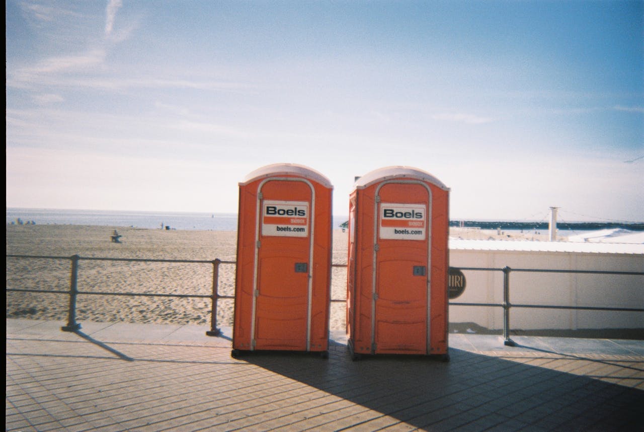 Home Two orange portable toilets on a beach promenade with blue sky and sand.