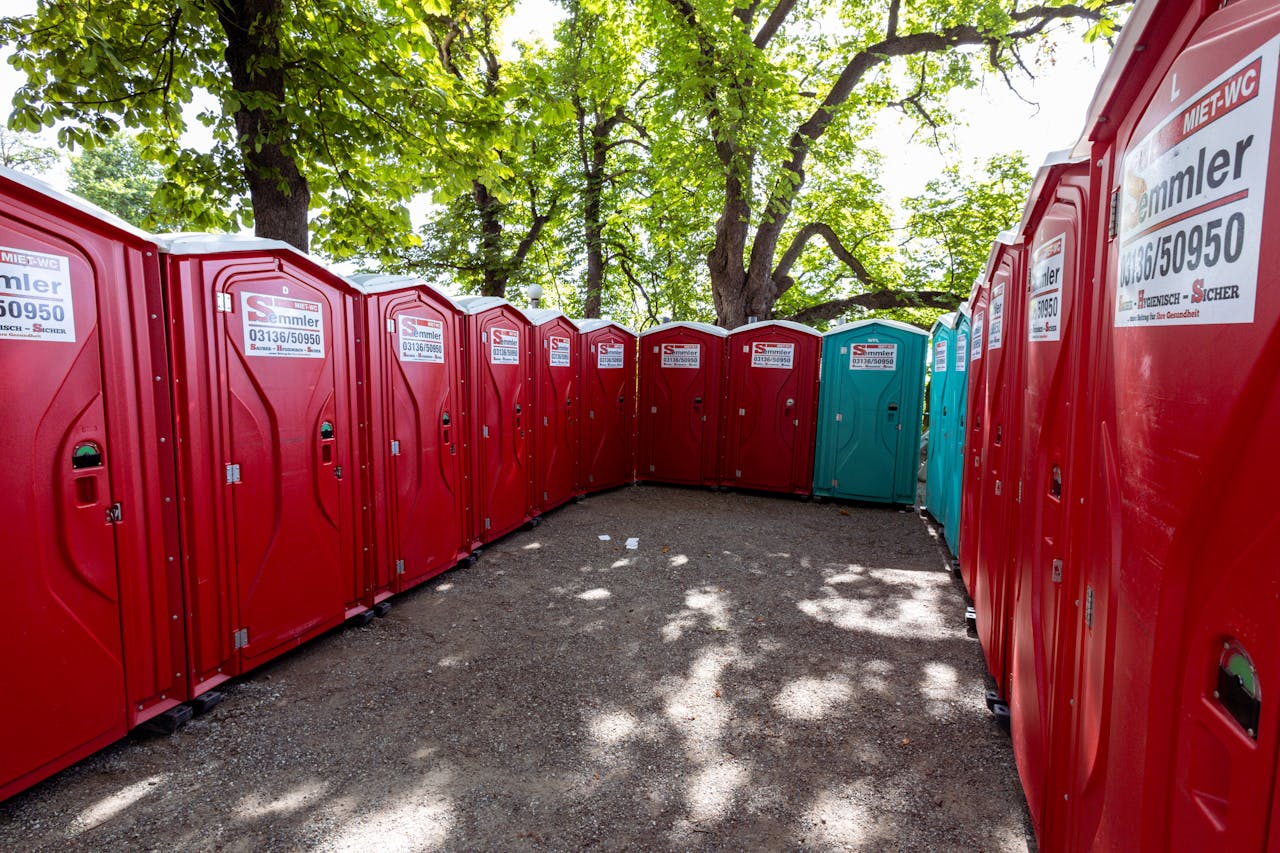 Services A line of red portable toilets under green trees in a sunny outdoor setting.