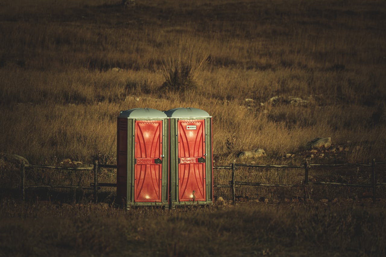 Services Two red portable toilets set in a vast, serene grassland during the day, capturing an unusual yet peaceful contrast.